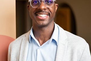 Evans Rochaste, NP, smiling while wearing clear glasses, a light gray blazer, and a blue button-down