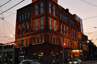 A Romanesque Revival style building at dusk at a major intersection with cars passing through.