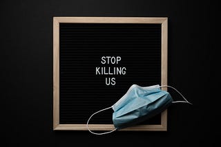 A black board with light wood frame displaying white lettering that says: “STOP KILLING US” in all capitals. There’s a medical face mask in the bottom right hand corner.