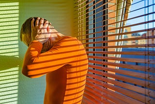 Distressed man with hands on head wearing an orange long-sleeved shirt standing by a window with venetian blinds