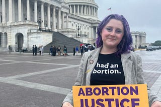 A photo of Lizzie, a White woman with purple hair. She is wearing a T-shirt that reads “ had an abortion,” and holding a sign that says “ABORTION JUSTICE NOW. The Capital building is in the background.