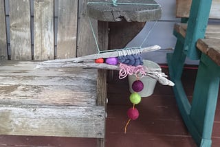 An odd little driftwood split-branch weaving with felted wool beads hanging from the arm of an old, weathered Adirondack chair