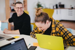 Two people sitting at a work desk in front of their laptops laughing.