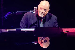 Billy Joel sitting a piano, smiling slightly and looking to the side. His face is reflected in the piano’s surface.