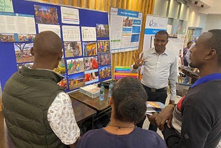 A man wearing a white shirt holds up his hand while standing beside a poster showing implementation of field work. There are three people standing in the foreground with their backs to the camera, taking in the presentation.