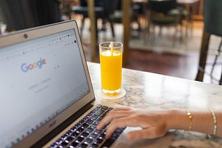 A woman types something on her laptop, but you can only see her arm and not her face. There is a glass of orange juice beside the laptop, and the screen shows she is searching Google.