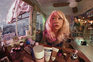 A still from the piece “Kowloon Forest.” A person sits at a table covered in makeup. They are illuminated in a pink light by a neon sign outside a large nearby window.