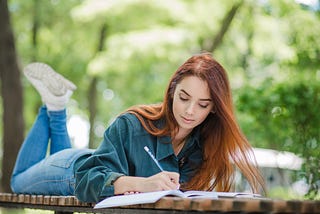 A girl lying on table in park writing.