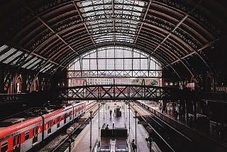 A train station that has a red train stopped on the left side, and some people waiting to board.