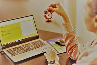 a woman looking at a tiny clock while sitting at a desk in front of her open laptop