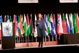 Image of a woman speaking on stage with the flags of all nations behind her and a banner that says, Rotary Connects the World.