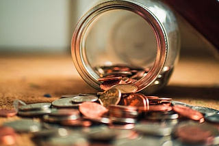 A glass jar tipped over spilling coins onto a flat surface. The view is looking directly inside the tipped over jar