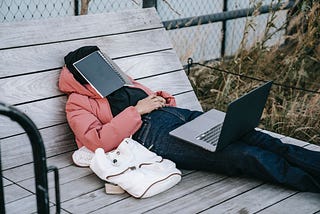 Exhausted person lying outside, on a large wooden bench, with a laptop on their lap and a notebook over their face.