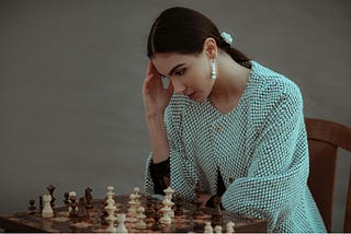A young woman in a light blue knitted sweater sits at a wooden chess board, resting her head in her hand in a contemplative pose. She wears pearl drop earrings and has her dark hair pulled back with a light blue hair accessory. The chess pieces are set up on an ornate wooden board, with both light and dark pieces visible. The lighting is soft and the background is neutral, creating an intimate, thoughtful atmosphere.