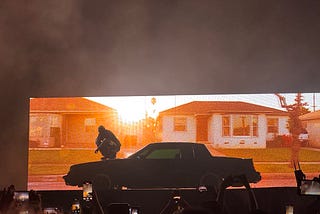 Kendrick lamar crouching on the hood of his Buick GNX while rapping into a microphone.
