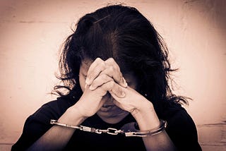 A dark-haired woman leaning her against her handcuffed and clasped hands.