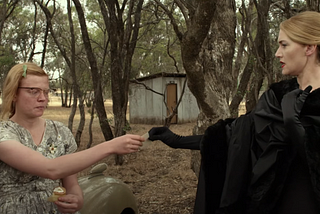 A screencap from The Dressmaker, showing Tilly in haute couture handing her calling card to the plainly-dressed Gertrude.