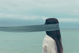 person facing the right with a wide teal ribbon covering their face and trailing far behind them out of the image, person’s head is covered in long, straight dark hair falling onto a white shirt, opaque sea or lake and cloudy sky in the background