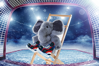 A stuffed elephant on a chair, wearing hockey stakes, in a hockey net