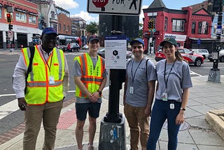 Four District Department of Transportation team members standing by a DTPR sign near a crosswalk at a busy intersection. The sign uses the DTPR visual language to provide notice of and describe the technology being tested, and a QR code enables residents to get more information and provide their feedback.