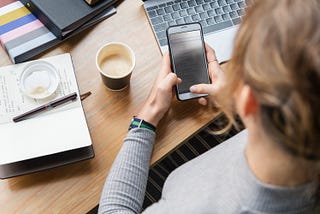 Person holding a mobile phone at a desk with a journal and coffee.