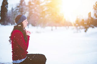 A woman in a red sweater praying and meditating, with the sun shining in the background over a wintery landscape.