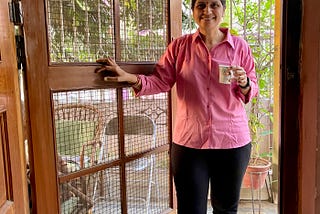A woman wearing a pink, full-sleeved shirt with a collar and black trousers is holding a coffee cup and is smiling at the camera.