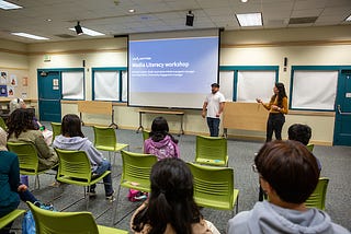 Ana Clara Otoni and Michael Lozano stand in front of a crowd of a dozen of teens in a library room presenting a slide show where it reads: “Media literacy workshop.”