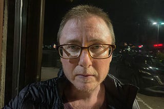 The author is a white man with short hair wearing glasses. He has a serious expression. It’s dark outside. A streetlight has lit up his face. He’s wearing a black jacket. He has stubble of a beard and mustache.