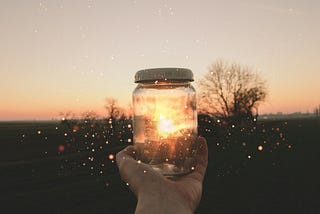 Hand holding a clear jar containing fireflies with a background of the sun setting.