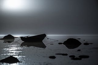 Moody black and white photo. A smooshed blob in the sky is the moon. In dark silhouette on the surface of water is a small boat tied to several large rocks.