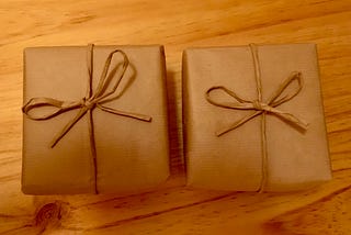 Two small gifts wrapped in brown paper and tied with brown paper strings into bows, sitting on a wooden surface.