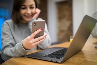 Mulher segura um celular enquanto esta em frente a um notebook