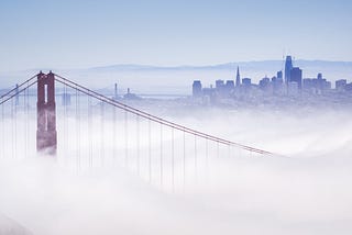 Photo of fog overflowing the Golden Gate Bridge and invading the city. You can see one tower of the bridge in muted red, with cables sweeping down and away from it, artistically, and disappearing into the fluffy white. In the background is the bluish charcoal grey skyline of San Francisco. The sky is a dull blue getting ready for twilight. You might think of the fog as a blanket, but it’s chilly rather than cozy.