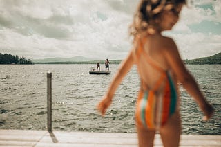Child in a striped bathing suit standing by a lake in the foreground with two older people standing on a floating raft in the background.