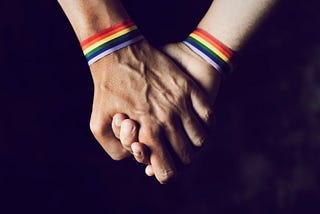 Two white men wearing rainbow wristbands holding hands over a black background