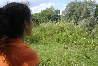 Woman in orange t-shirt looking at lush grassy field and blue sky.