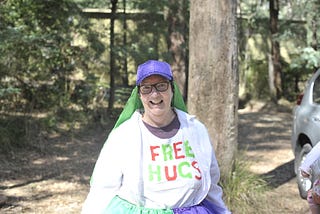 A woman in a purple sparkly hat and black-framed glasses smiles at the camera. She wears a white t-shirt that says FREE HUGS.