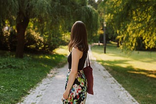 A woman walking alone on a narrow path surrounded by trees, with her back to the camera.