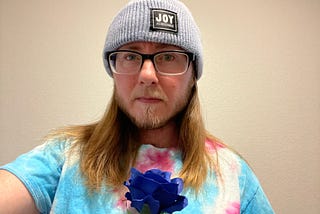 The author is wearing a knitted blue hat reading “Joy as Resistance”, black rimmed glasses, and a blue, white, and pink shirt. He is holding a dark blue flower. He has a beard and mustache and a stern look with a crease between his eyebrows.