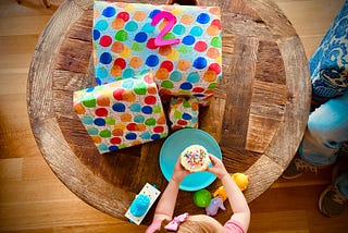 photo shows a table with a pile of presents wrapped in balloon wrapping paper with the number 2 on top. In front of the gifts, a pair of small arms reach over some discarded toys to hold a vanilla cupcake with sprinkles