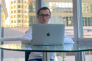A man in his mid 20s wearing a white sweather and blue jeans. He’s sitting on a chair. He’s resting a MacBook against a glass table. He has black hair and glasses. Behind him is a window and several skyscrapers.