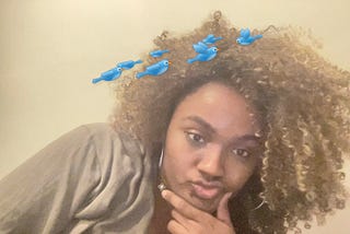 A black woman with a blonde fro looks into the camera with a funny face. Fake birds swirl over her head.