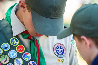 A Boy Scout in a baseball cap looks down at a sash covered in colorful merit badges as another boy points to them.