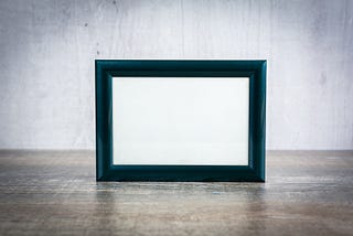 An empty picture frame by a white wall, standing on a wooden top.
