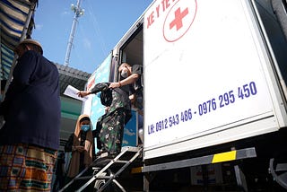 A woman walks down a short flight of steps outside of a mobile TB clinic with support from one of the health workers leaning outside of the mobile van.