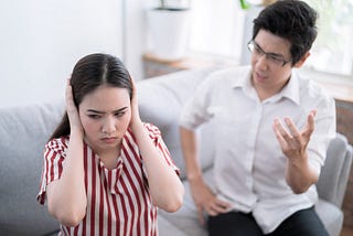 An Asian couple sitting on a sofa, clearly in the middle of a difficult discussion. The man has his hand raised, trying to argue his point, while the woman has her hands over her ears, trying not to listen to what he is saying.