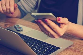 A white femme person holds a mobile phone in front of her open laptop.