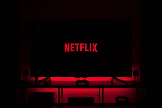 An image of the Netflix logo on a television screen in a dark room. The television is on top of a cabinet with a console gaming controller sitting in front of it. The red from the logo font resonates outward from the television giving a sort of ominous glow.