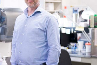 Robert Kozak, in a blue shirt, hands in pants’ pockets, stands by the sink of his research laboratory.